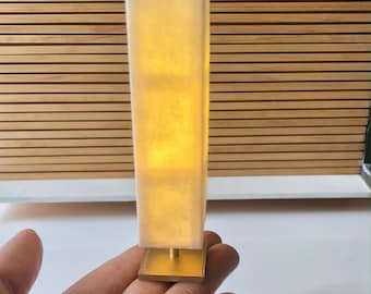 Op de afbeelding: Een witte lamp met een gouden voet. De lamp is aan en geeft een warm licht.