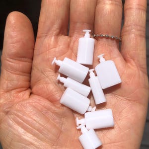 Puede incluir: Una colección de siete pequeños frascos de plástico blanco con tapas de bomba. Los frascos están dispuestos en una pila en la mano de una persona.