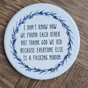 May include: A circular white coaster with a blue floral wreath. The text on the coaster reads: "I DON'T KNOW HOW WE FOUND EACH OTHER BUT THANK GOD WE DID BECAUSE EVERYONE ELSE IS A FUCKING MORON."