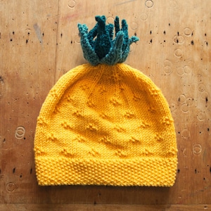 Op de afbeelding: Een gele gebreide muts met een groene en blauwe pompon bovenop. De muts heeft een textuurpatroon en is gemaakt van zacht garen.