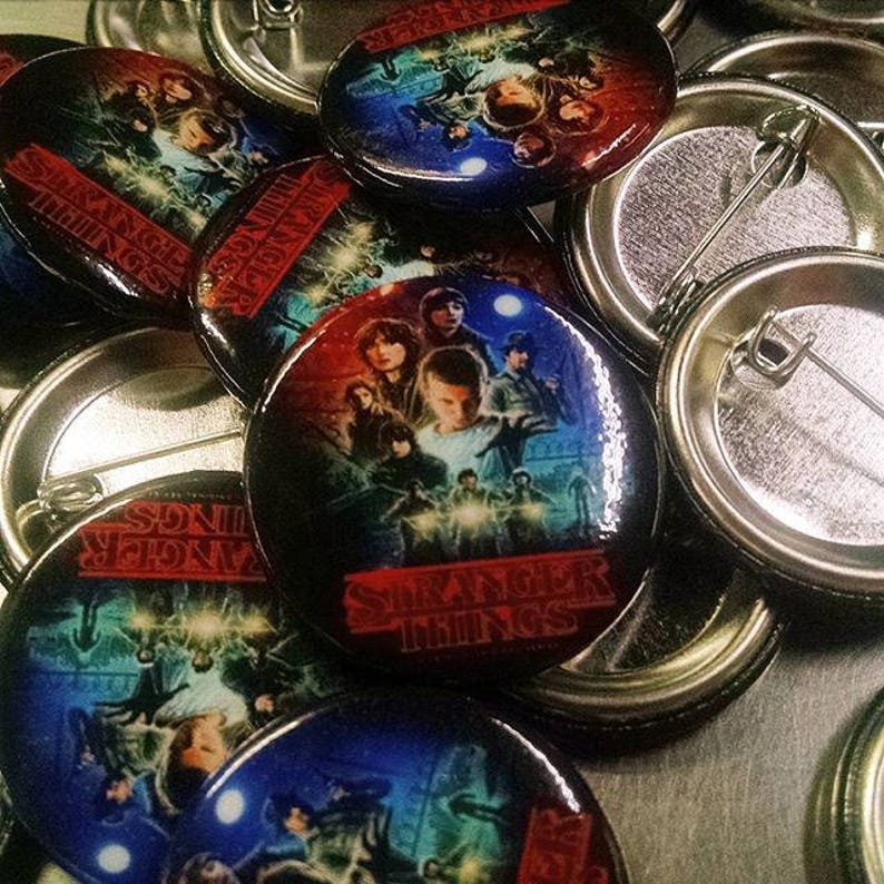 May include: A collection of round, metal pins with colorful images of the characters from the show "Stranger Things". The pins feature the show's logo and the text "Stranger Things".