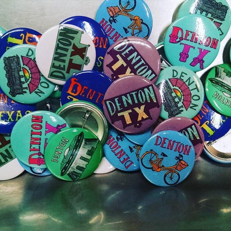 May include: A collection of colorful round buttons with the text "Denton TX" on them. Some buttons feature a train, a bicycle, and a UFO design.