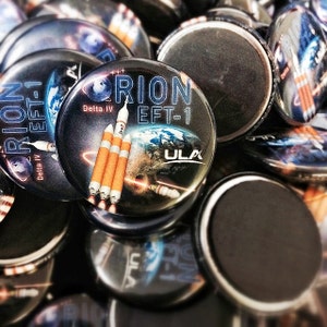 May include: Close-up of a collection of round, black and white buttons with the text "Orion EFT-1" and "Delta IV" printed on them. The buttons feature a graphic of a rocket launch with the text "ULA" printed on them.