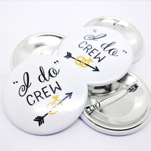 May include: White round button with black text that reads "I do Crew" with a gold ring and arrow design. The button has a silver back with a safety pin.