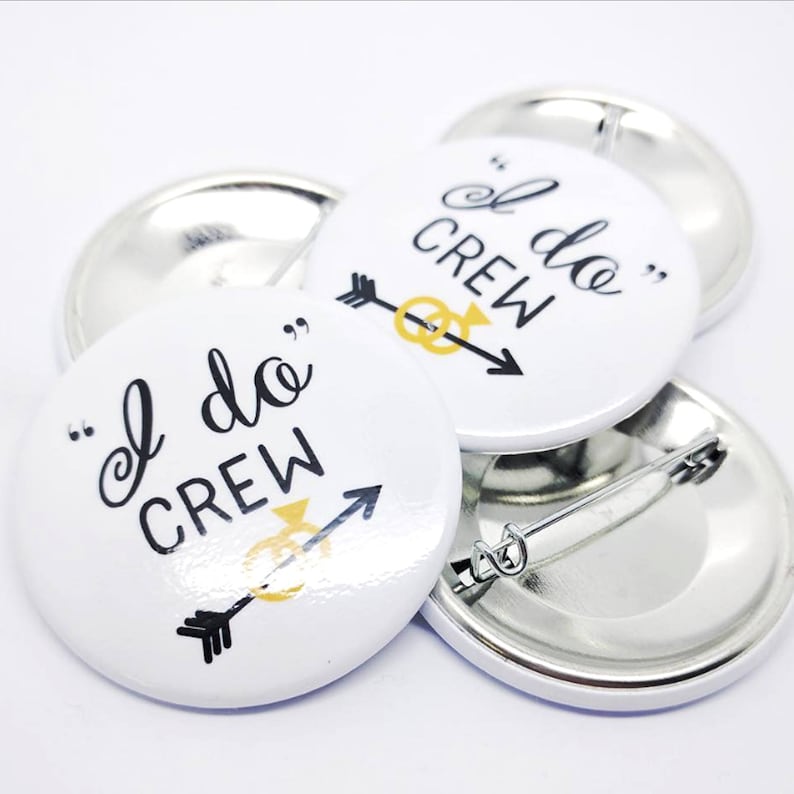 May include: White round button with black text that reads "I do Crew" with a gold ring and arrow design. The button has a silver back with a safety pin.