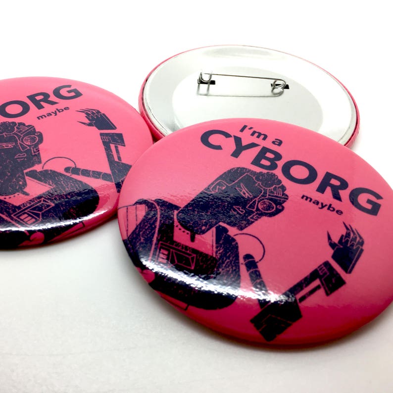 May include: Two pink button pins with a black and white illustration of a person with a robotic arm. The text on the pins reads "I'm a CYBORG maybe".