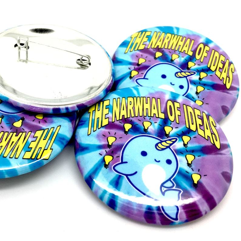 May include: Three round button pins with a tie-dye background. The buttons feature a cartoon narwhal with a yellow horn and light blue body. The text "The Narwhal of Ideas" is printed in yellow on the buttons. The buttons have a silver metal backing with a safety pin.