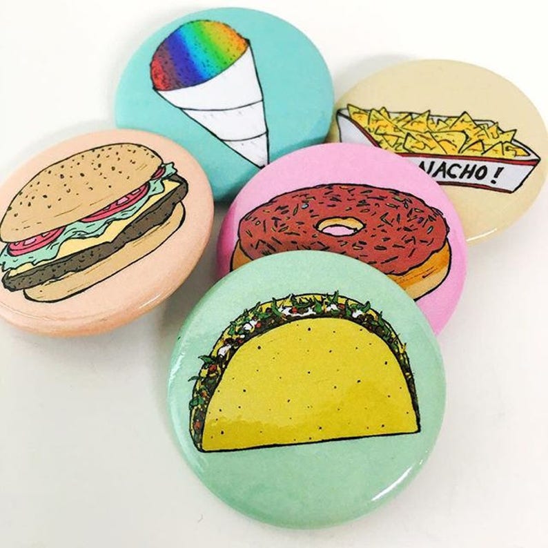 May include: Five colorful round pins with different food illustrations. The pins feature a hamburger, a donut, a taco, a snow cone, and nachos. The pins are all about 2 inches in diameter.