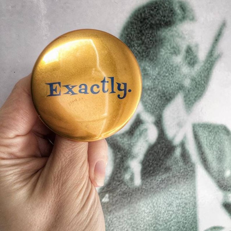 May include: A gold-colored round button with the word "Exactly." printed in blue.