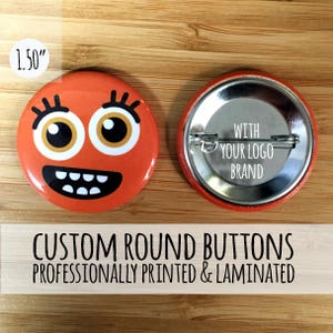 May include: Two 1.5 inch round custom buttons. One button features a cartoon face with orange skin, black eyes, and a wide smile. The other button is blank with the text "WITH YOUR LOGO BRAND".