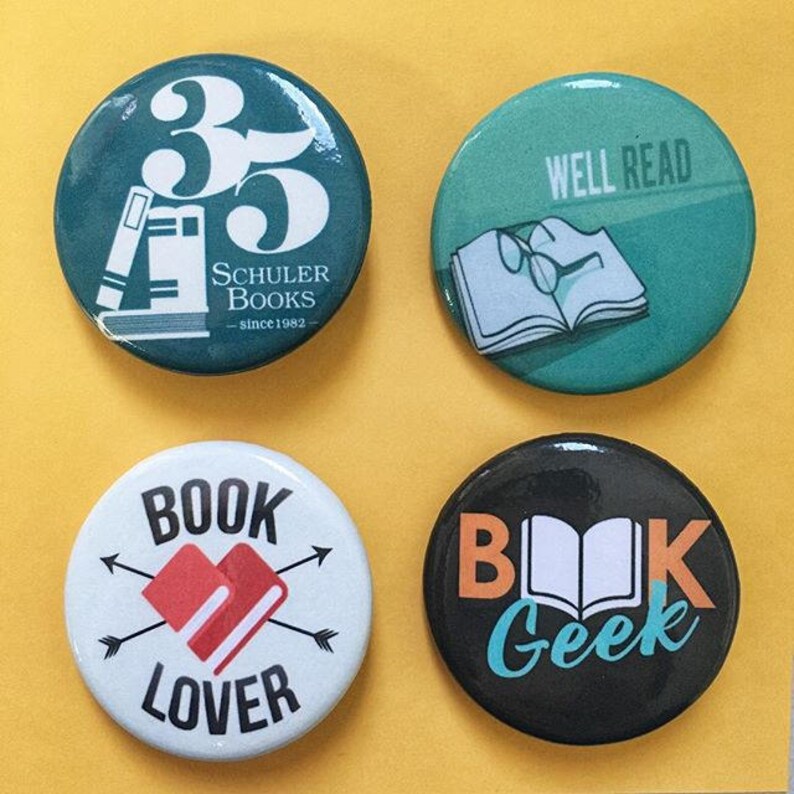 May include: Four round button pins with different designs. The first pin is teal green with white text that says "35" and "Schuler Books - since 1982". The second pin is teal green with white text that says "Well Read" and a white open book with glasses on top. The third pin is white with red text that says "Book Lover" and a red heart with a book inside. The fourth pin is black with orange text that says "Book Geek" and a white open book.
