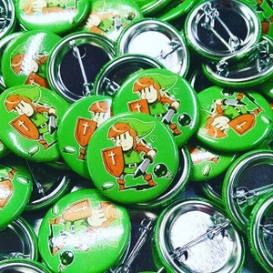 May include: A close-up of a pile of green button pins featuring a cartoon character with a sword and shield. The character is wearing a green hat and a green tunic. The buttons are all the same size and have a silver back.