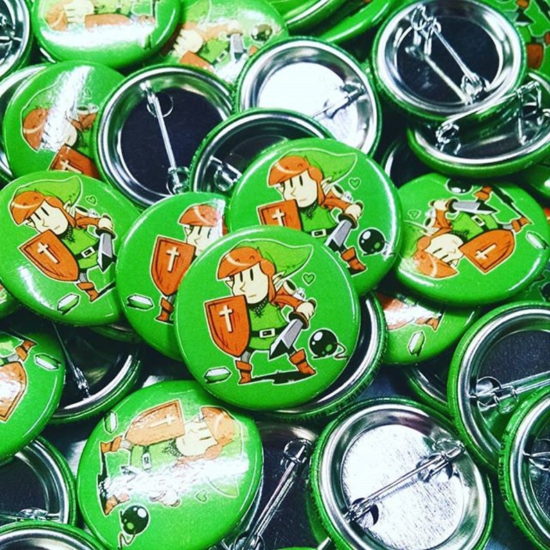 May include: A close-up of a pile of green button pins featuring a cartoon character with a sword and shield. The character is wearing a green hat and a green tunic. The buttons are all the same size and have a silver back.