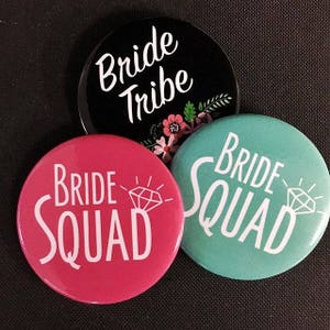 May include: Three round buttons with the text "Bride Tribe", "Bride Squad" and "Bride Squad" in white lettering. The buttons are black, pink and teal.