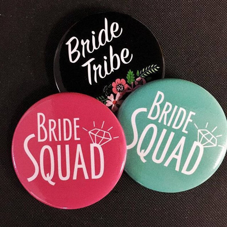 May include: Three round buttons with the text "Bride Tribe", "Bride Squad" and "Bride Squad" in white lettering. The buttons are black, pink and teal.