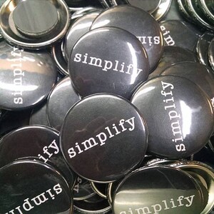 May include: A close-up of a pile of silver-colored round buttons with the word "simplify" printed in white on each button.
