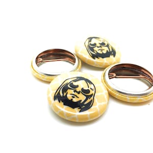 May include: Four yellow button pins with a black and white image of a person wearing sunglasses. The pins have a honeycomb pattern.