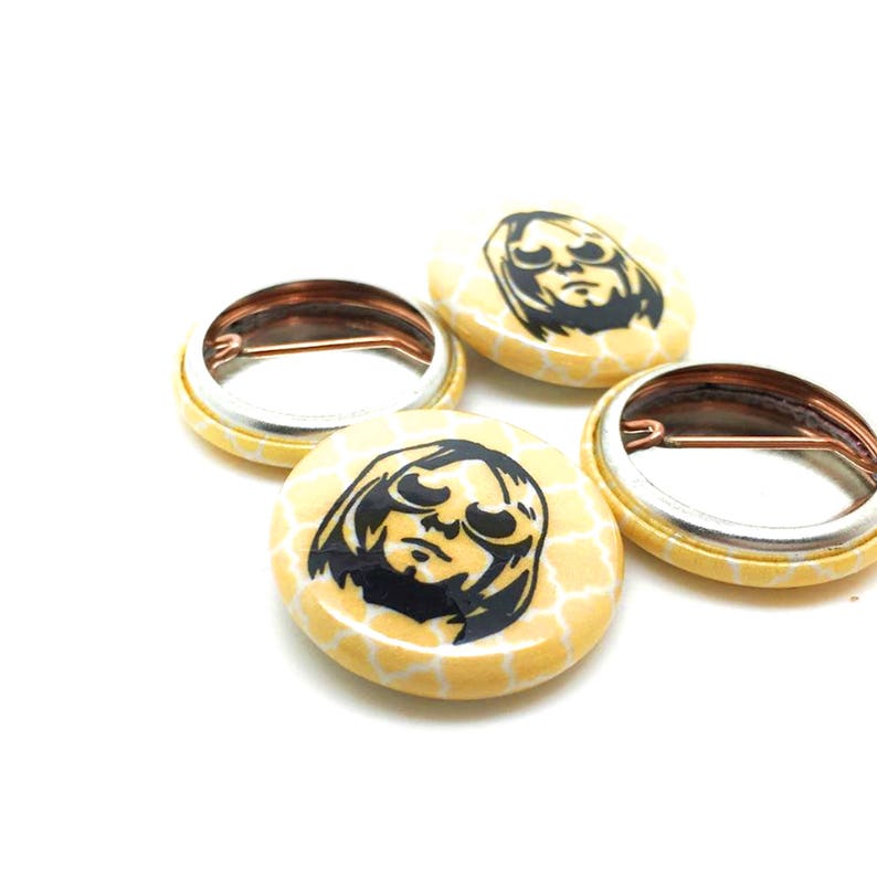 May include: Four yellow button pins with a black and white image of a person wearing sunglasses. The pins have a honeycomb pattern.