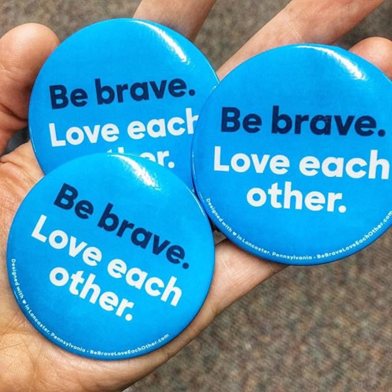 May include: Three blue round buttons with the text "Be brave. Love each other." in white lettering. The buttons are designed with a heart in Lancaster, Pennsylvania. The website address BeBraveLoveEachOther.com is printed on the bottom of each button.