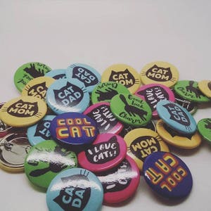 May include: A collection of colorful round pinback buttons with various cat-themed designs and text, including "Cool Cat", "Cat Dad", "Cat Mom", "I Love Cats!" and "Good Luck Charm".