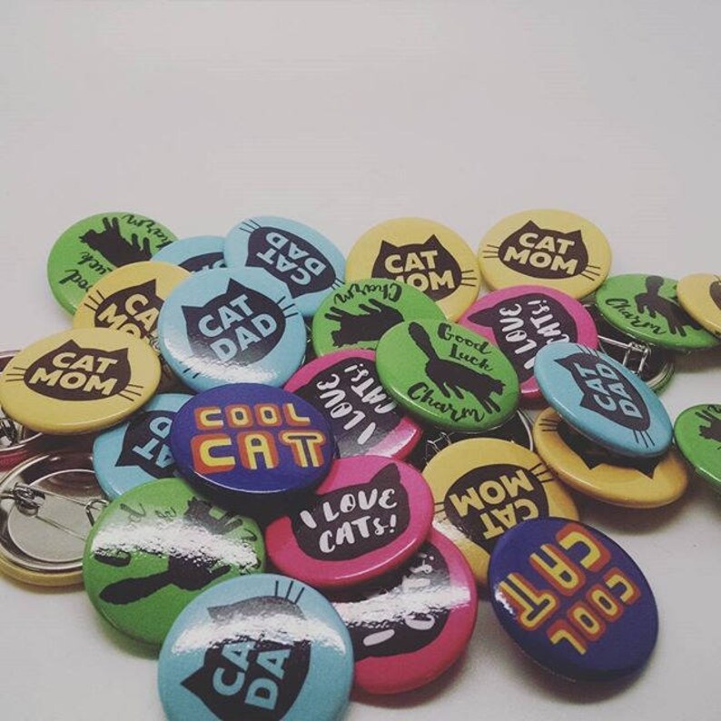 May include: A collection of colorful round pinback buttons with various cat-themed designs and text, including "Cool Cat", "Cat Dad", "Cat Mom", "I Love Cats!" and "Good Luck Charm".