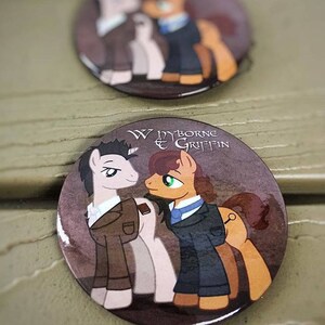 May include: Two cartoon ponies wearing suits. The pony on the left is gray and the pony on the right is brown. The text "Wyborne & Griffin" is printed on the button.