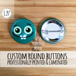 May include: A 1.25 inch round button with a green background and a cartoon face with a toothy grin. The back of the button is silver with a pin and a green border. The text "WITH YOUR LOGO BRAND" is printed below the button. The text "CUSTOM ROUND BUTTONS PROFESSIONALLY PRINTED & LAMINATED" is printed below the buttons.