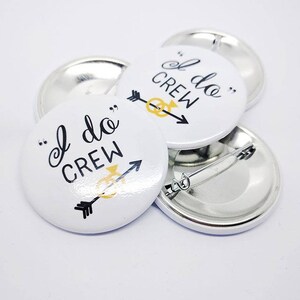 May include: Four white round buttons with a black and gold design. The buttons say "I do Crew" with an arrow and a gold ring. The buttons are attached to silver metal backs with safety pins.