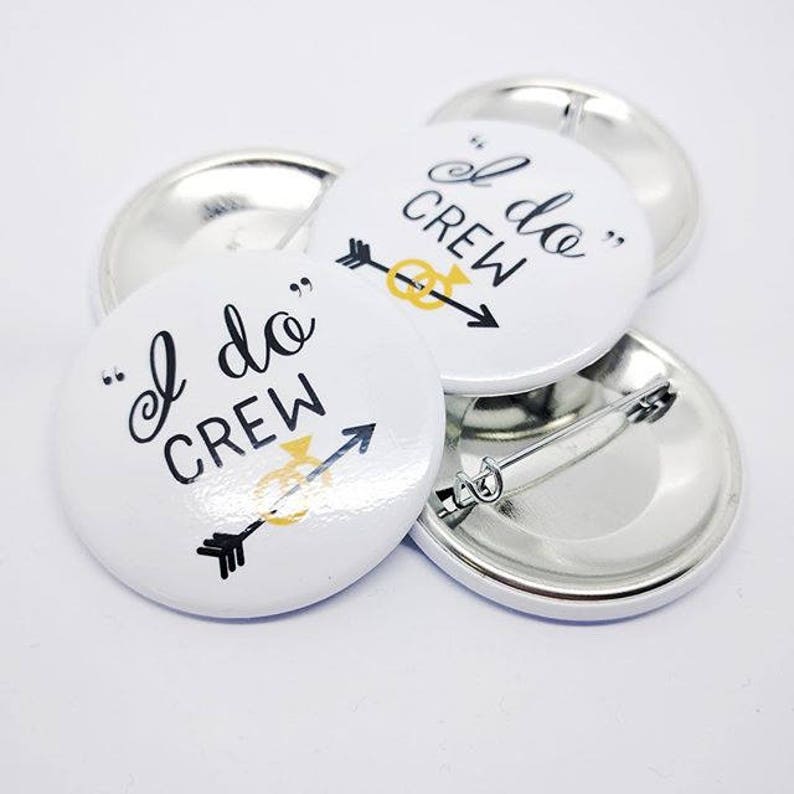 May include: Four white round buttons with a black and gold design. The buttons say "I do Crew" with an arrow and a gold ring. The buttons are attached to silver metal backs with safety pins.