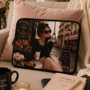 May include: Black laptop sleeve featuring a Parisian cafe scene with a woman in sunglasses, the Eiffel Tower, and the text "BOULANGERIE DE PARIS". A black mug with "CAFÉ DE PARIS" is visible. A pink pillow and a book titled "PARIS mon amour" are also in the image.