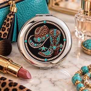 May include: A round silver compact mirror featuring a black center inlay with a graphic print of a leopard print cowboy hat and a matching cowboy boot, both embellished with turquoise-colored stones and clear crystals, framed by a crystal border.