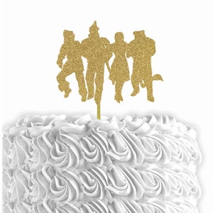 May include: Gold glitter cake topper featuring the silhouette of the characters from the Wizard of Oz. The characters are Dorothy, the Scarecrow, the Tin Man, and the Cowardly Lion.