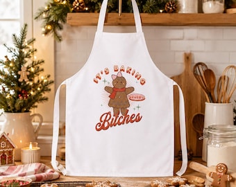 Puede incluir: Un delantal blanco con un gráfico central de una figura de pan de jengibre marrón con un lazo rosa y una bufanda roja, sosteniendo una bandeja. El texto dice "IT'S BAKING" en fuente roja arqueada y "Bitches" en letra cursiva roja grande.