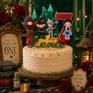 May include: A white frosted cake with a Wizard of Oz theme, featuring character toppers and the name "Lexa". The cake is decorated with green sprinkles and sits on a wooden cake stand. A sign reads "Happy Birthday Lexa!"