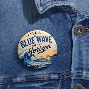 May include: A round button with the text "I SEE A BLUE WAVE ON THE HORIZON" in blue and white. The button features a graphic of a blue ocean wave with a setting sun in the background. The button is pinned to a denim jacket.