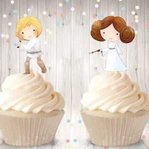 May include: Two cupcakes with white frosting and a Star Wars theme. The cupcake on the left has a cartoon image of Luke Skywalker holding a lightsaber. The cupcake on the right has a cartoon image of Princess Leia holding a blaster.