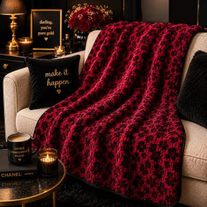 Op de afbeelding: Een rood-zwarte patroon deken over een beige bank. Een zwart kussen met de tekst "make it happen" ligt op de bank. Een goudkleurige lijst met de tekst "darling, you're pure gold" hangt aan de muur.