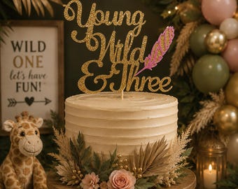 May include: A gold glitter cake topper featuring the text Young Wild & Three with a pink glitter feather accent. The topper is positioned on a beige frosted cake decorated with dried palm leaves, greenery, and small pink flowers.