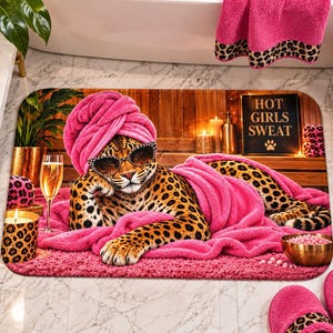 May include: A rectangular bath mat featuring a leopard wearing a bright pink towel wrap on its head and a matching pink blanket. The leopard wears leopard-print sunglasses. A sign in the background reads HOT GIRLS SWEAT with a paw print icon.