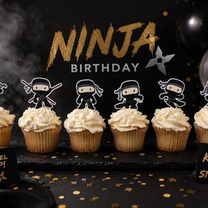 May include: A ninja-themed birthday display featuring cupcakes topped with ninja figures. The background includes the words "NINJA BIRTHDAY" in gold glitter, black balloons, and a silver shuriken. Two signs read "LEVEL UP!" and "BE KIND BE STRONG".