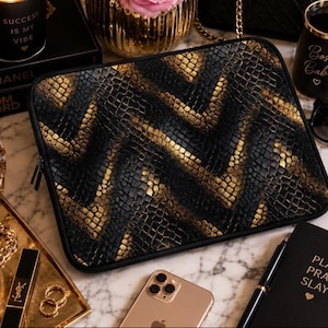 May include: A rectangular laptop sleeve featuring a textured black and gold snakeskin pattern arranged in a chevron design. The sleeve has rounded corners, a black fabric border, and a zipper closure along the top edge for securing a device inside.