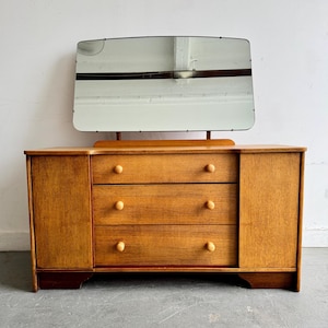 May include: A light brown wooden chest of drawers featuring three central drawers with round knobs, flanked by two side cabinets. A rectangular mirror with rounded corners is attached to the top, reflecting the ceiling and a horizontal structural beam.