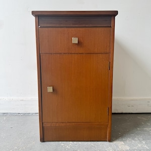 Puede incluir: Una mesita de noche de madera de color marrón medio con una superficie rectangular, un cajón superior y una puerta de armario inferior. Tanto el cajón como la puerta tienen pequeños pomos metálicos cuadrados y texturizados de color dorado.