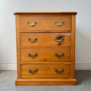 May include: A light brown wooden chest of drawers featuring four horizontal drawers. Each drawer is equipped with two brass swing-style handles. The wood grain is visible throughout the piece, and the structure sits on a flat base.