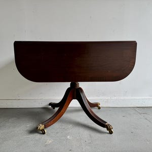 May include: An antique drop-leaf table in dark brown, featuring a round top and a pedestal base. The table has a curved edge and rests on three curved legs with brass castors. The table is made of wood and has a glossy finish. The table's dimensions are approximately 76 cm in height.