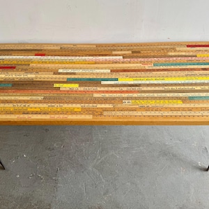 May include: A rectangular coffee table with a tabletop constructed from numerous vintage wooden rulers of varying lengths and colours, including yellow, red, and natural wood tones. The table is supported by four thin, black metal hairpin legs.