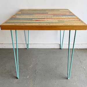 May include: A square table with a tabletop composed of various vintage wooden rulers arranged in horizontal rows. The table features four light blue metal hairpin legs and a natural wood frame edge. Text on the rulers includes ROC LIGHTING.