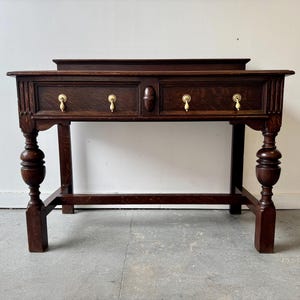 May include: A dark brown wooden console table with two drawers and brass handles. The table features turned legs and a rectangular top with a raised back. The drawers have decorative pulls.