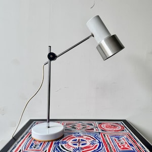 May include: A white and silver desk lamp with an adjustable arm and a round base. The lamp is positioned on a table with a colourful geometric pattern in red, blue, and black.