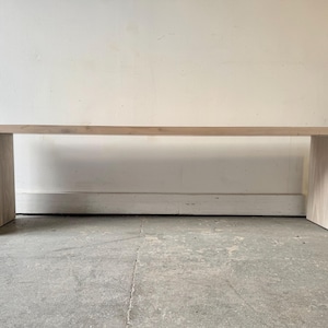 May include: A light-coloured wooden console table with a rectangular top and two vertical supports. The table is made of light-coloured wood and has a simple, modern design. The table is approximately 183 cm long.